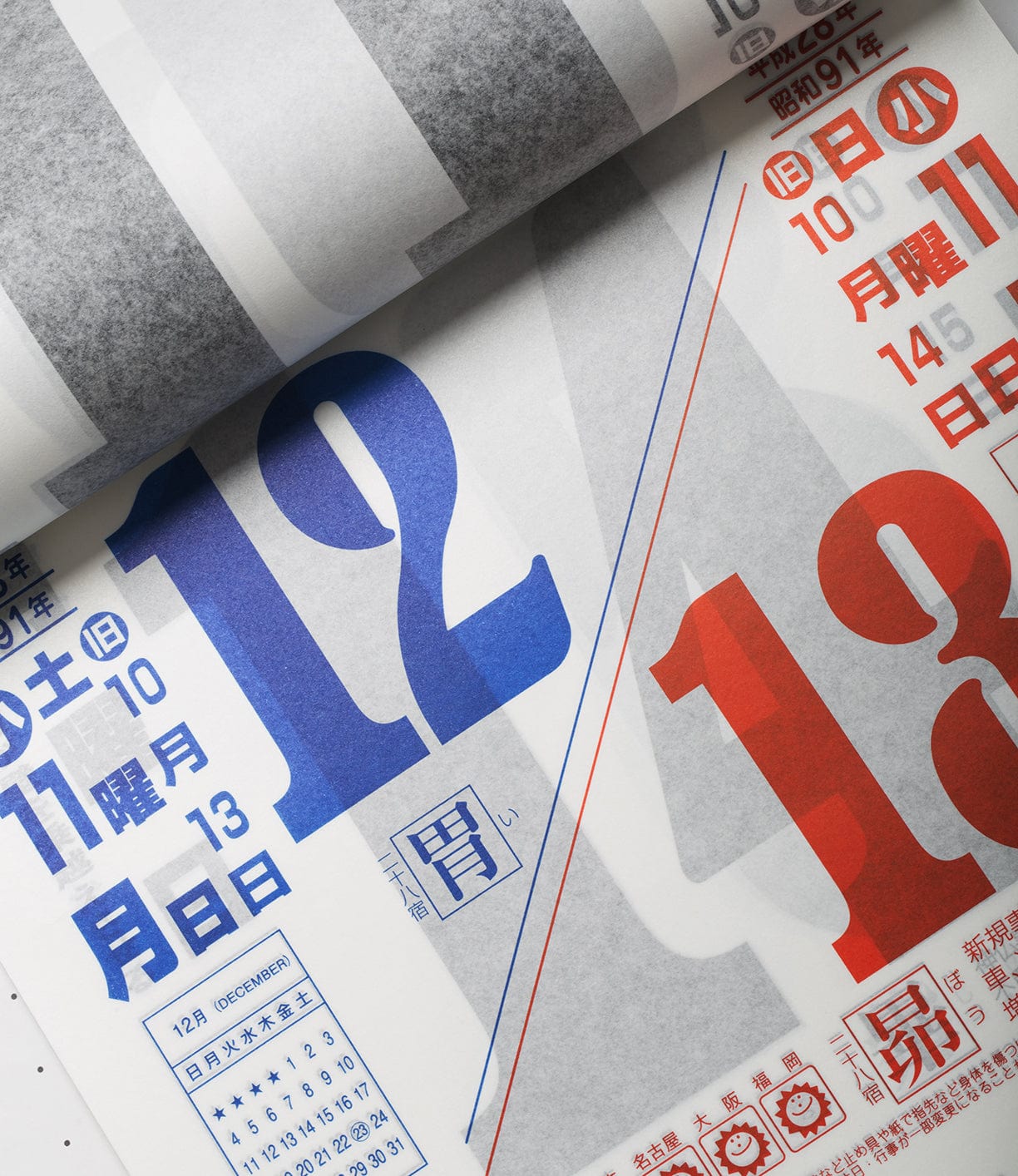 Close-up of a Japanese calendar, showing the dates 12 and 13 prominently in blue and red. The calendar text is in Japanese.