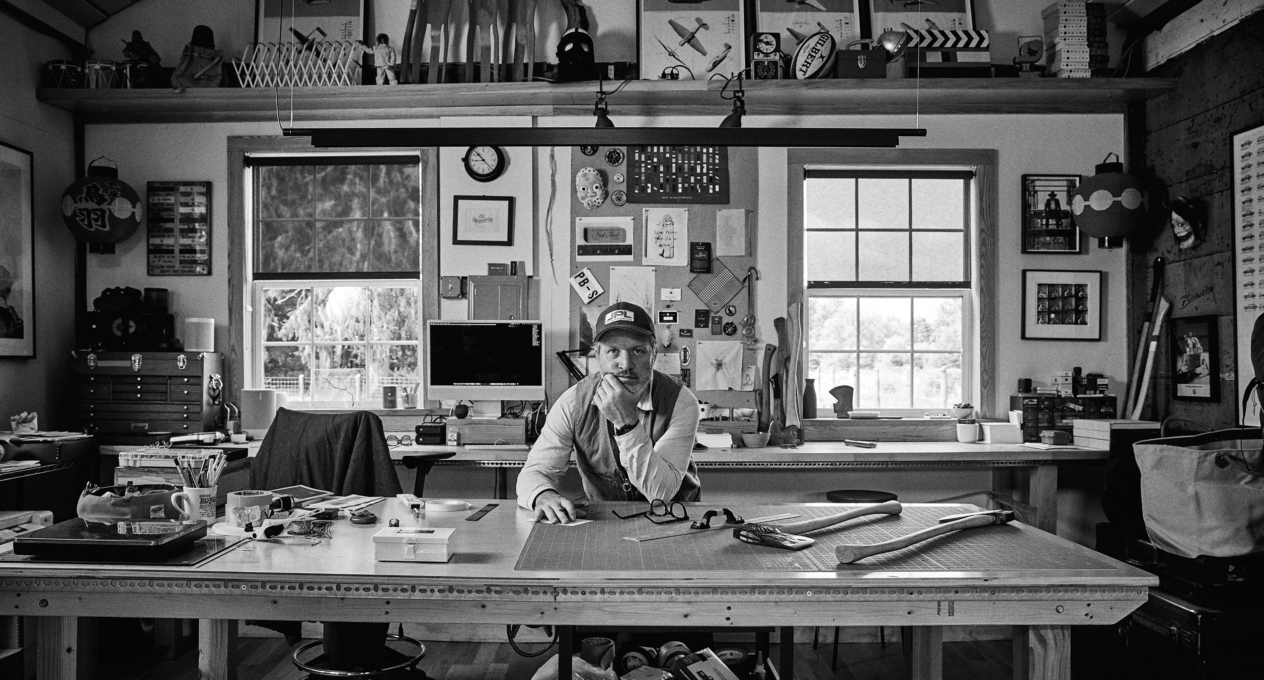 A person wearing a hat sits at a cluttered wooden desk in a workshop, surrounded by tools, papers, and various objects with large windows in the background.