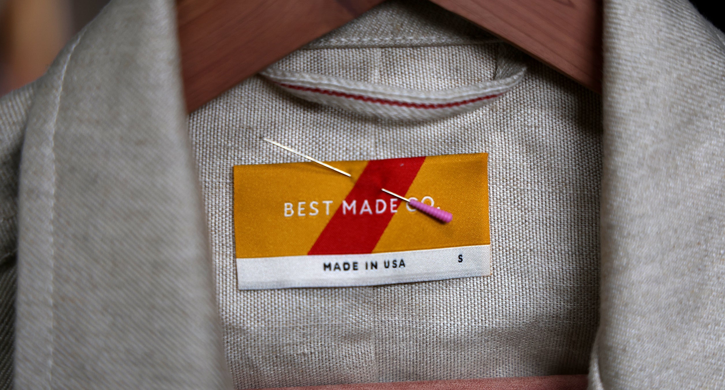 Close-up of a clothing label that reads Best Made Co., Made in USA, S with a red and yellow background. The label is attached to a beige garment on a hanger, with a needle and thread resting on it.