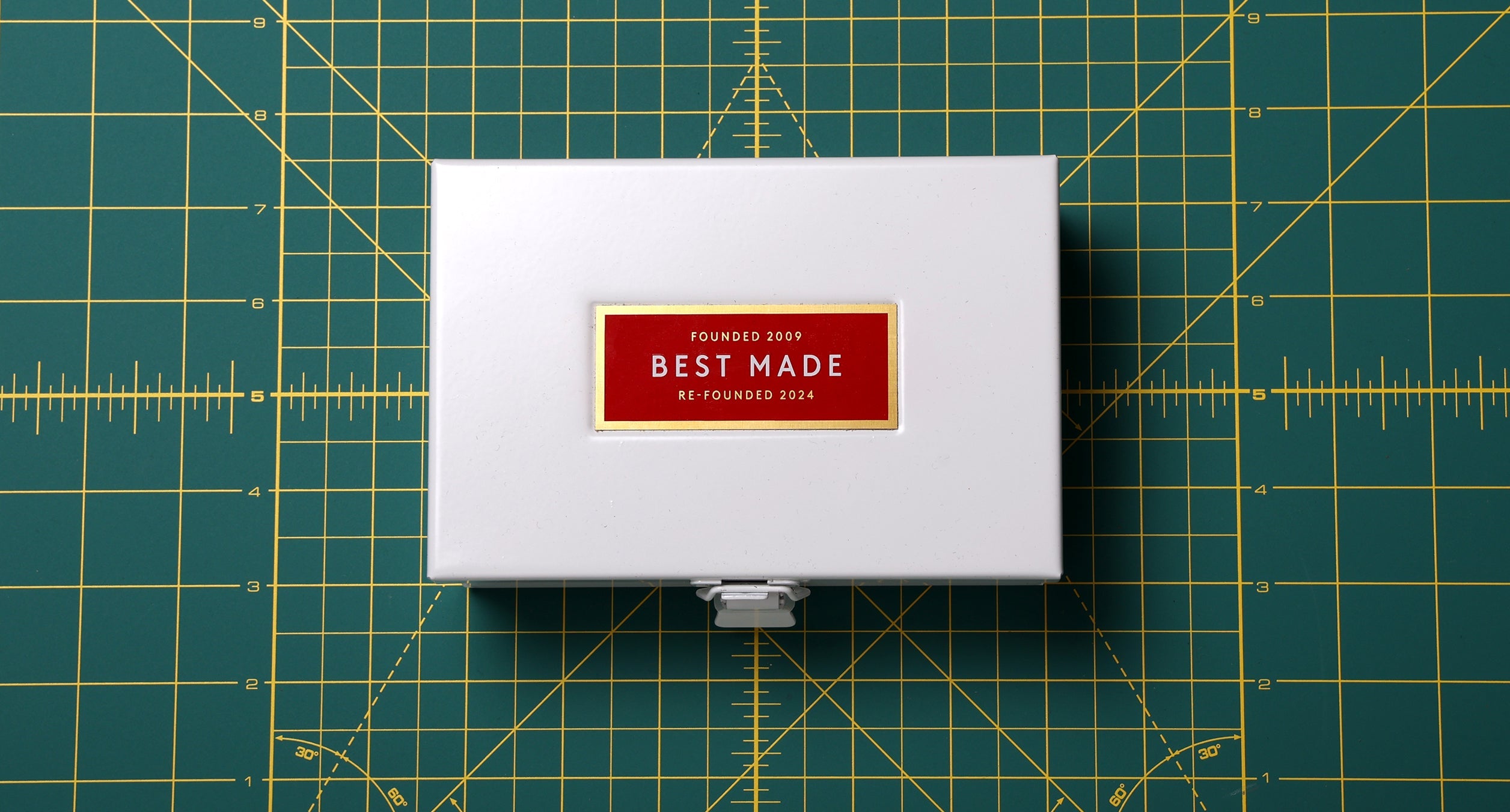 White rectangular box with a red label on top reading FOUNDED 2009 BEST MADE RE-FOUNDED 2024 is centered on a green cutting mat with yellow measurement lines.