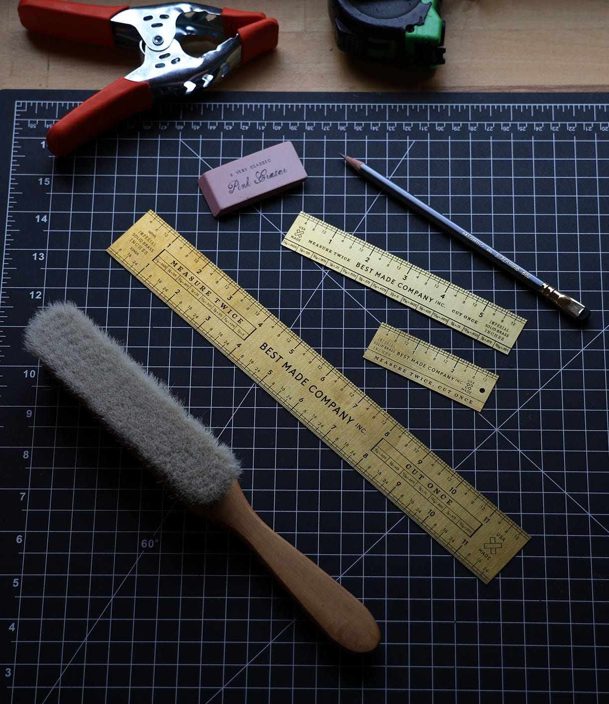 An overhead view of a cutting mat with a metal ruler, eraser, pencil, dust brush, hole punch, and measuring tape on it.