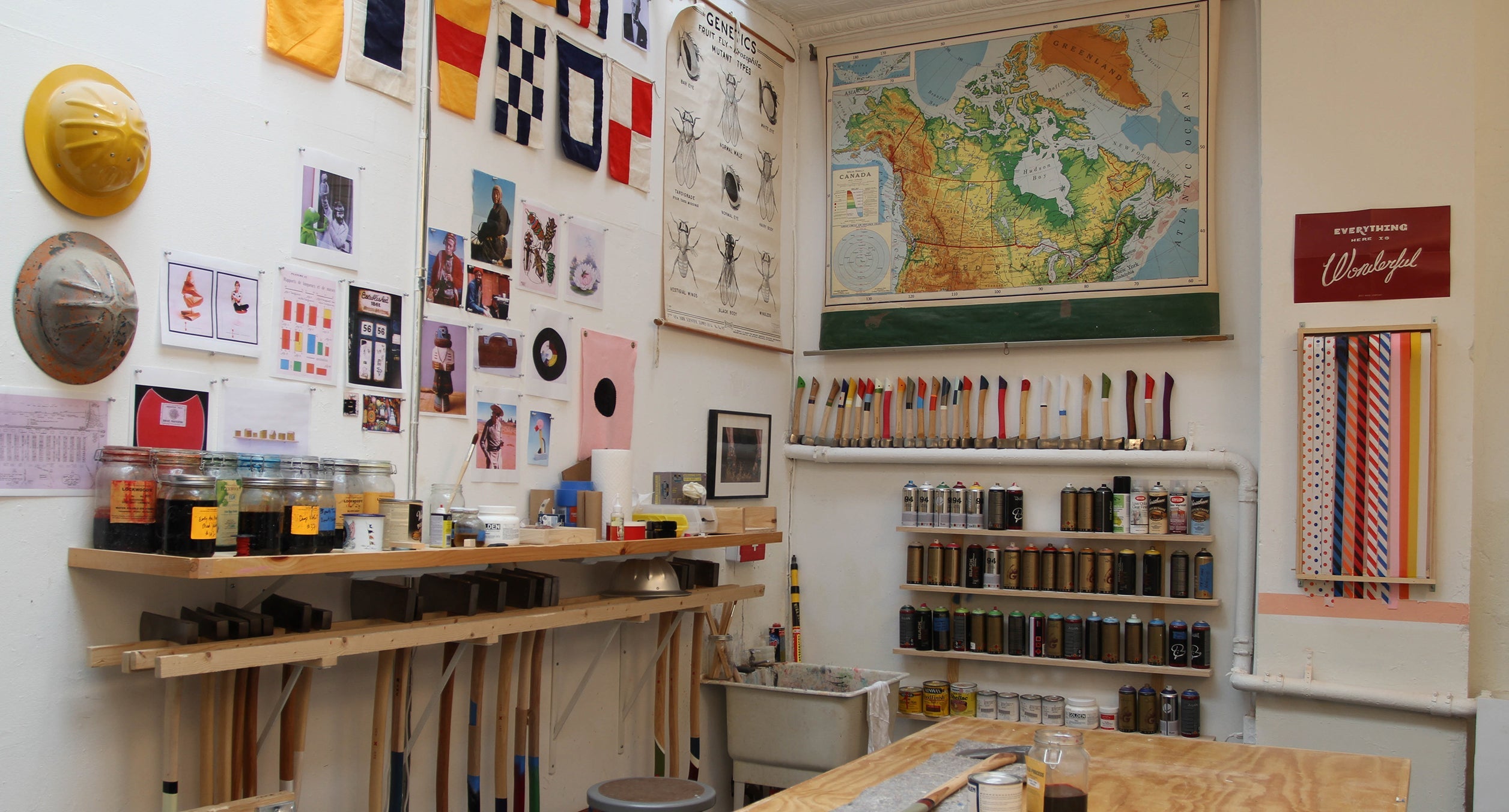 A workshop with nautical flags, a map, various tools, jars of materials, paint cans on shelves, and a rack of striped ribbons along the wall.