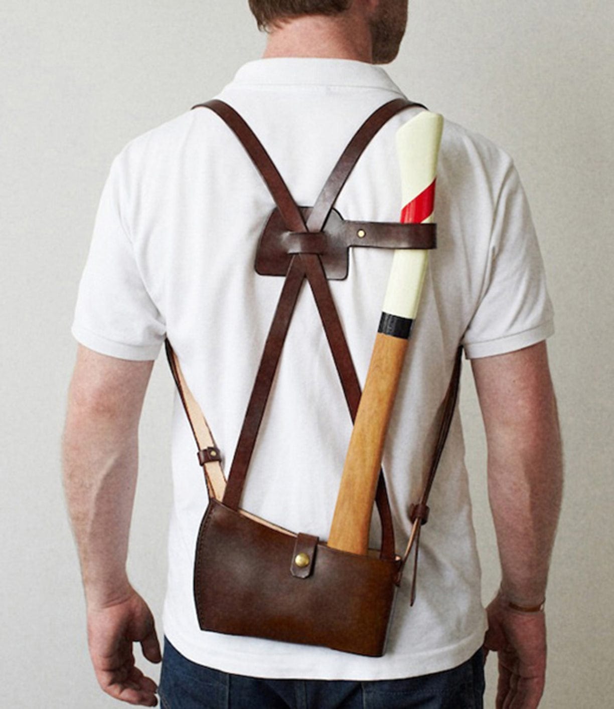 A person wearing a white shirt and blue jeans is seen from behind, equipped with a brown leather axe holster carrying a wooden axe with a white and red handle.