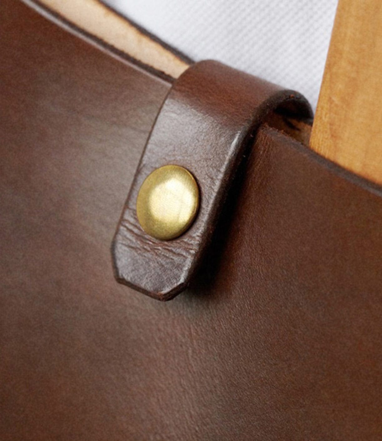 Close-up of a brass rivet on a brown leather strap loop, attached to a wooden handle on a white background.