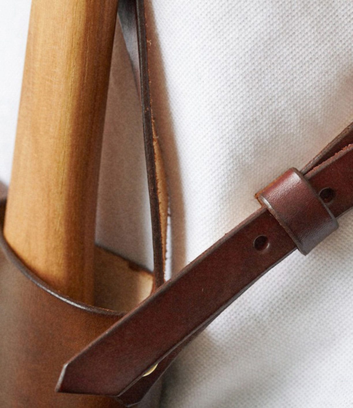 Close-up image of a wooden-handled tool, possibly an axe, secured in a brown leather sheath against a white fabric background.