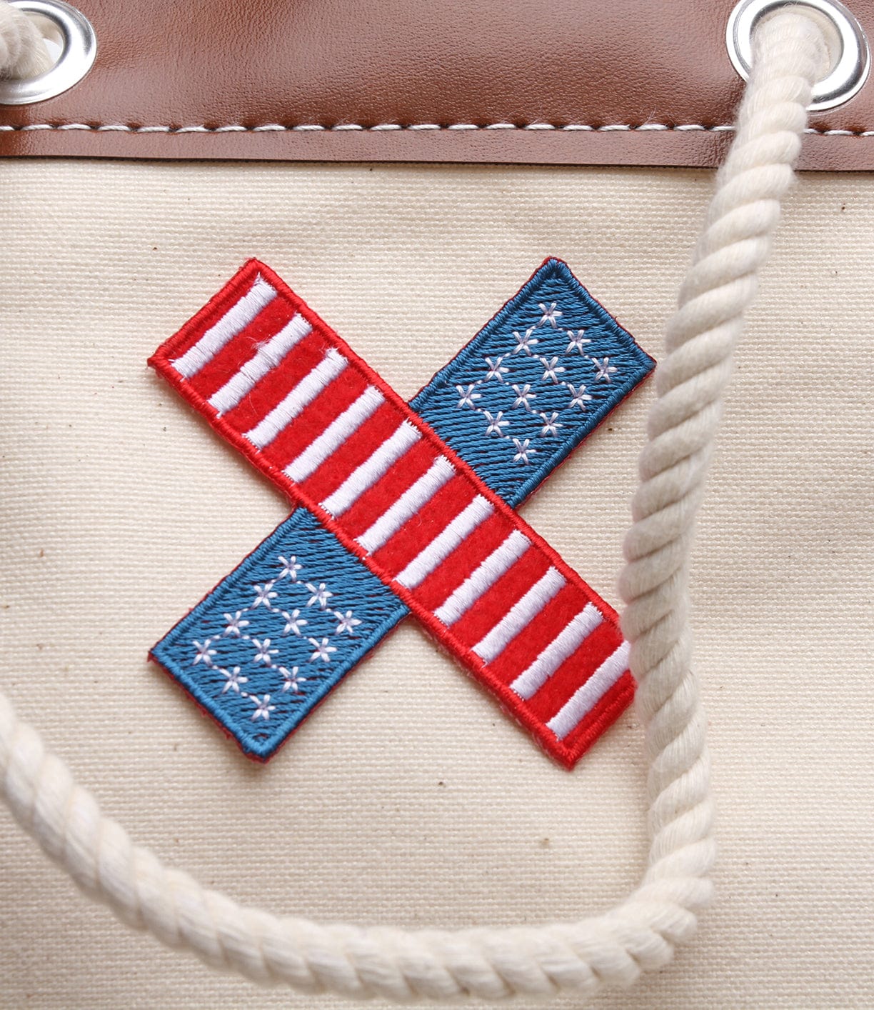 Close-up of a fabric patch featuring two crossed American flag motifs on a beige surface with white rope handles and brown leather trim.