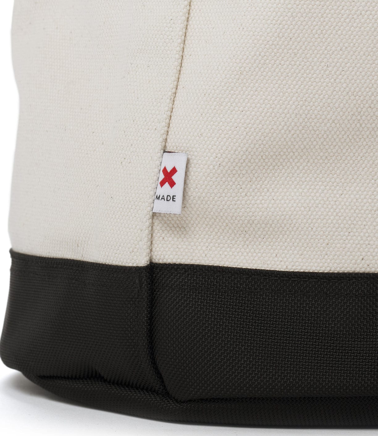 Close-up of a beige canvas bag with a dark green base and a small white tag featuring a red X and the word MADE stitched on the bag's side seam.