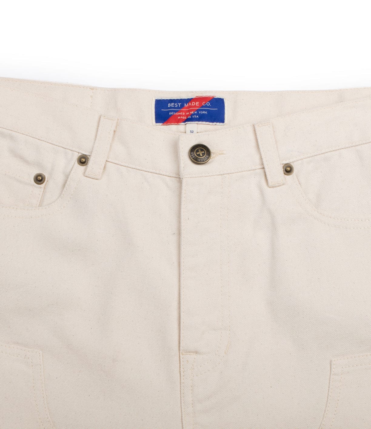 Close-up of a pair of beige pants with front pockets, button, and zipper. A blue label inside the waistband reads Best Made Co in white text with a red arrow design.