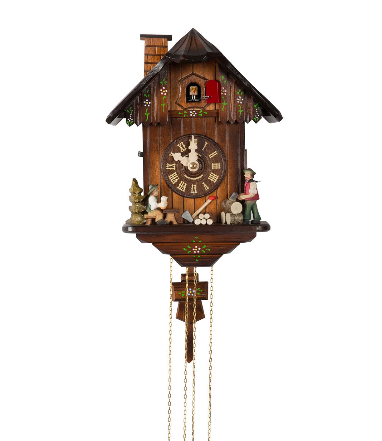 A wooden cuckoo clock with intricate carvings, featuring a small figure with an ax near a log pile, and a forest scene. The clock has Roman numerals and decorative chains hanging below.