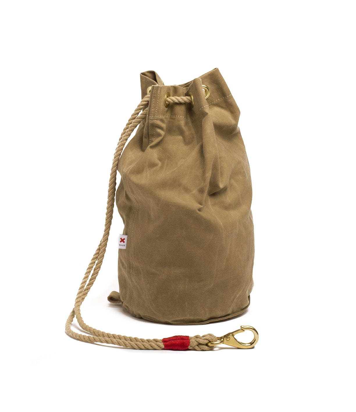 A tan canvas drawstring bag with beige rope handles, a side label featuring a small red cross, and a metal clip attached to the rope.