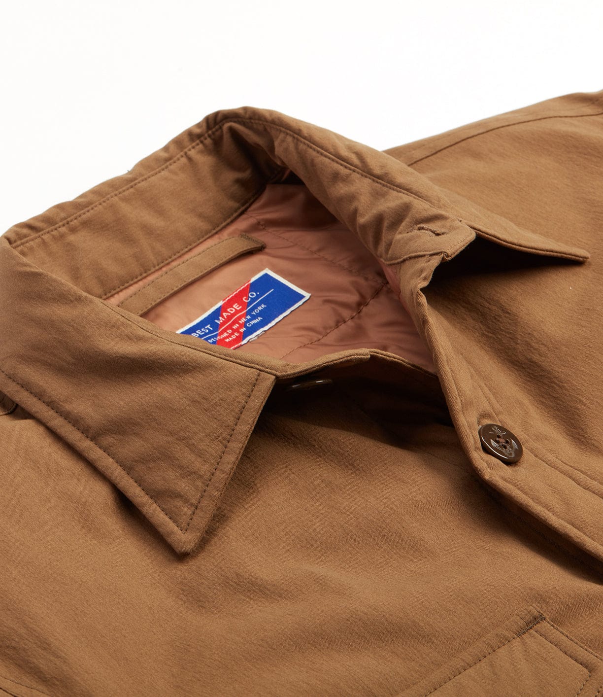 Close-up image of a brown collared jacket with a blue and red label visible inside. The jacket has a buttoned front and appears to be made of a sturdy fabric.