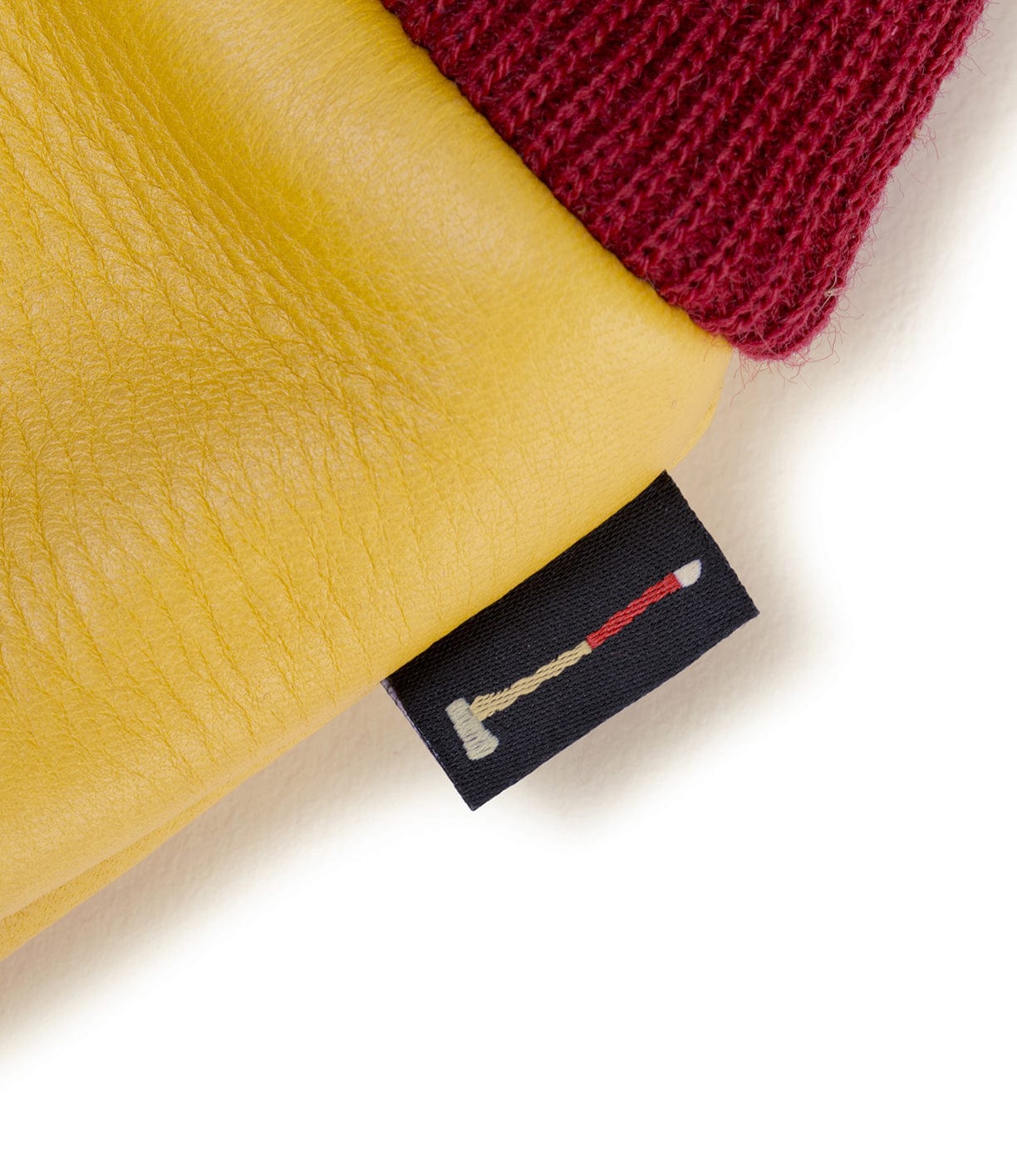 Close-up of a yellow leather item, possibly a glove, with a maroon knitted edge and a small black tag displaying a matchstick graphic.