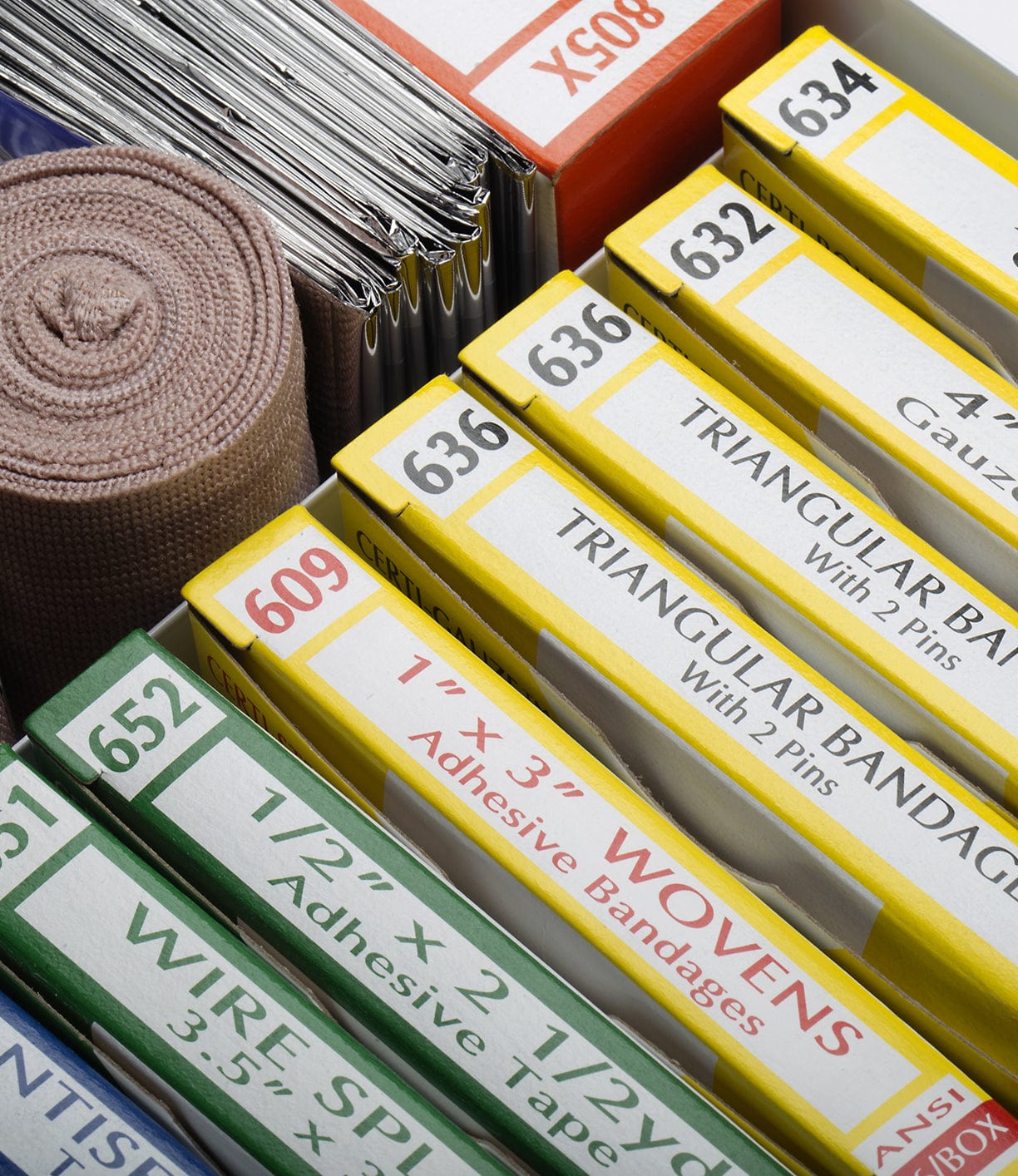 Close-up of various medical supplies, including gauze pads, triangular bandages, woven adhesive bandages, wire splints, and elastic bandage, organized in a drawer.