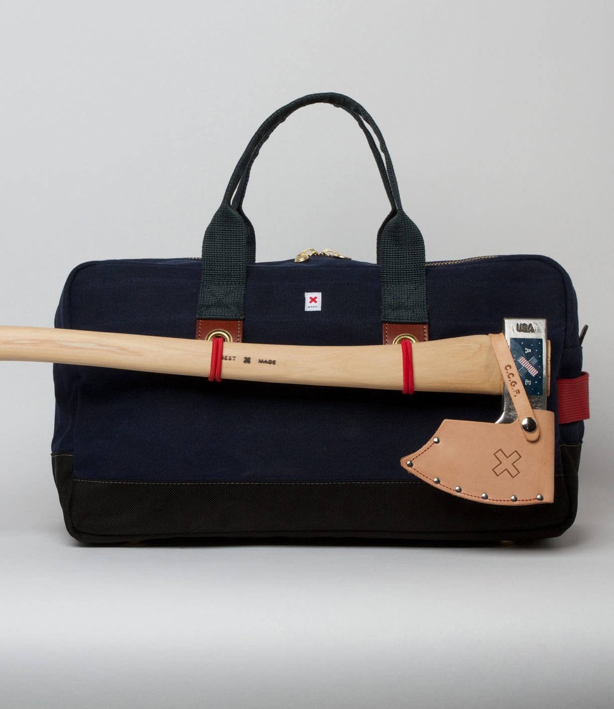 A navy and black duffel bag with a leather-handled axe attached to the front, secured by red straps.