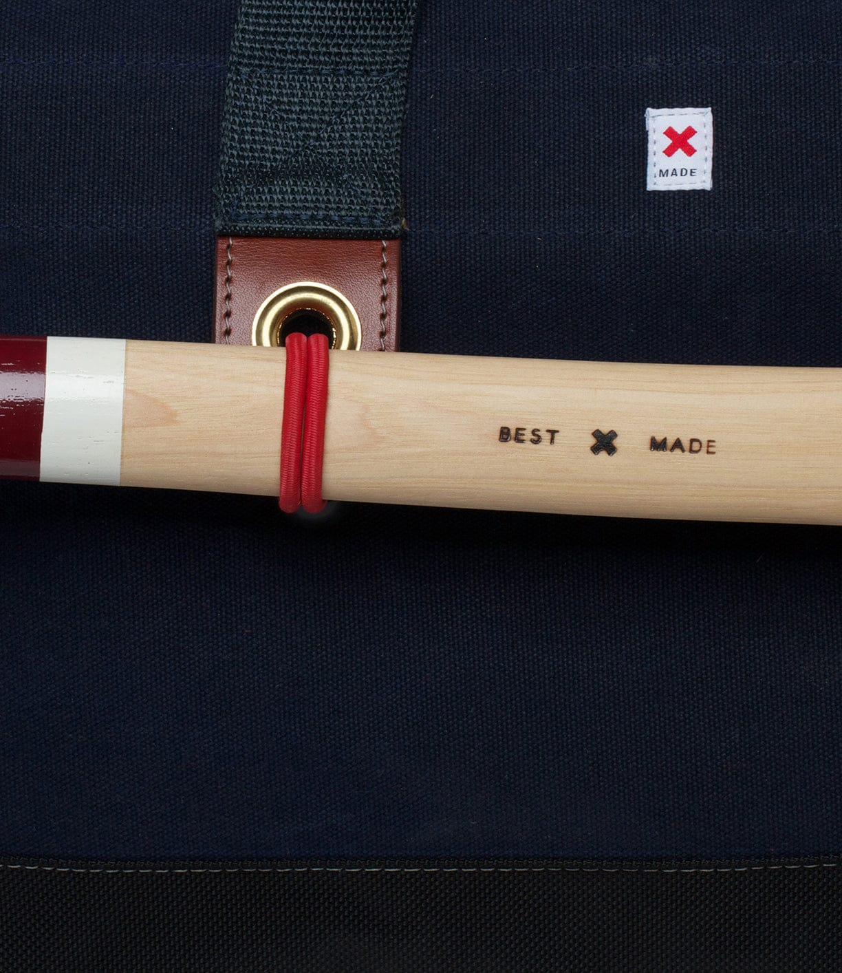 Close-up of a wooden handle marked with BEST MADE secured by a red band to a dark fabric background with a small white label featuring a red X logo.