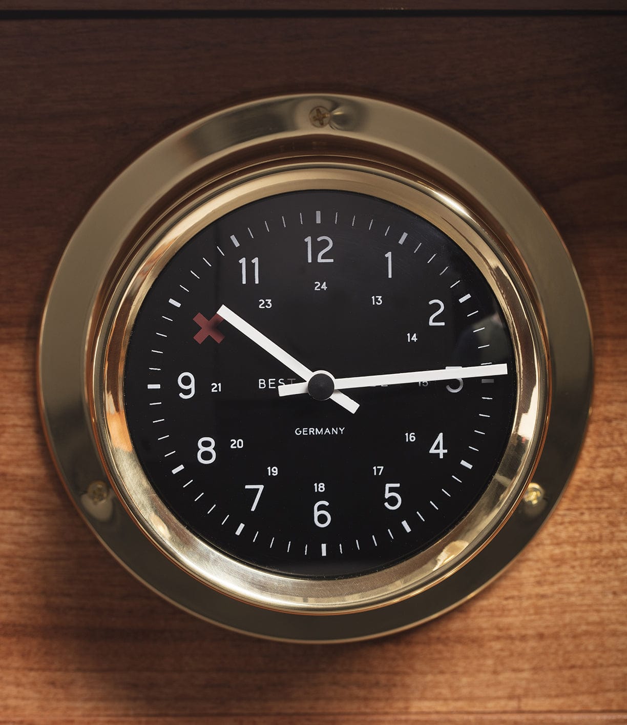 A round, black wall clock with a gold frame, featuring both 12-hour and 24-hour time formats, and labeled Bespoke Germany positioned against a wooden backdrop.