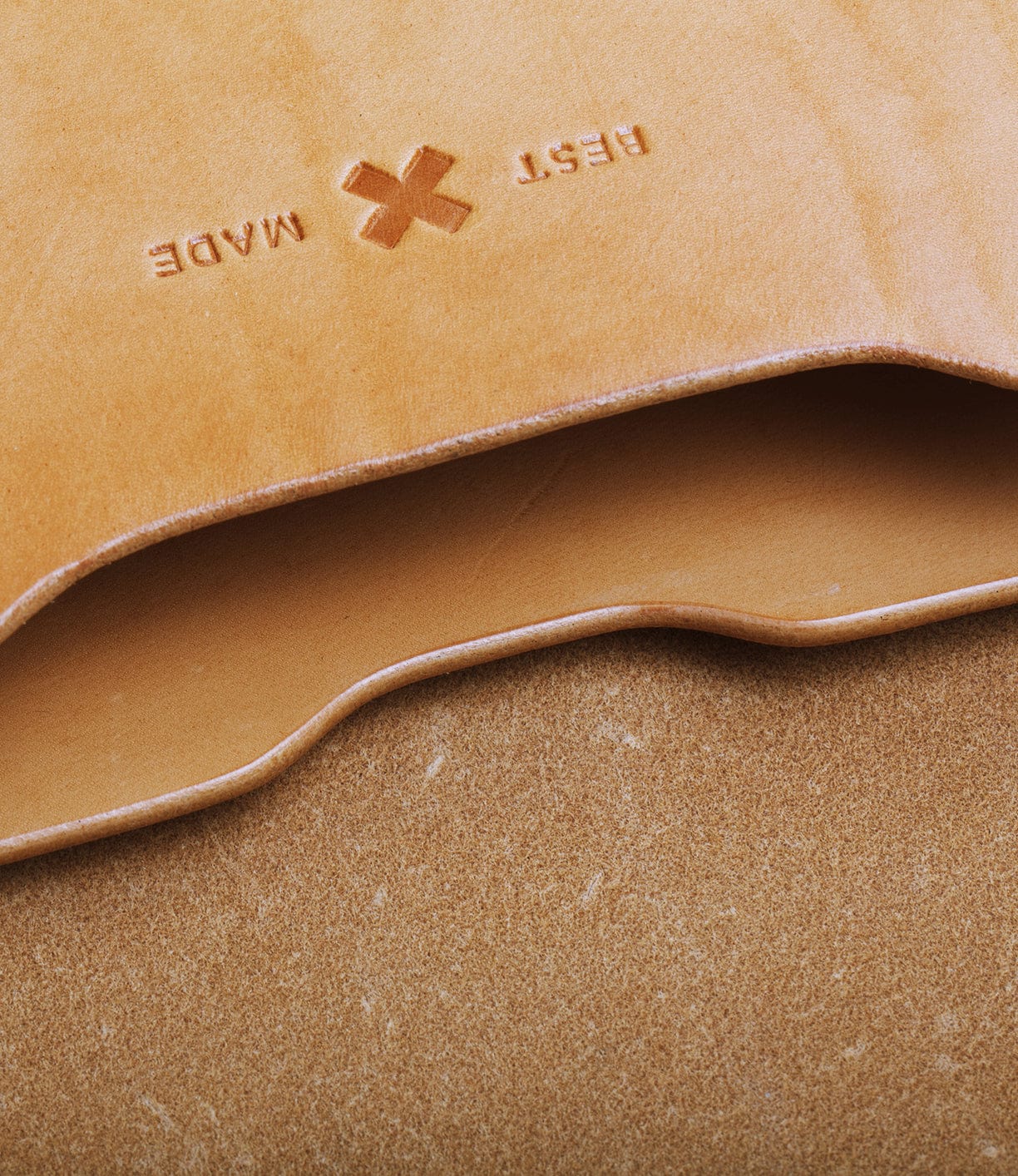 Close-up of a tan leather piece with a debossed BEST MADE and an X symbol. Edges are shown, showcasing the leather's texture and natural imperfections.