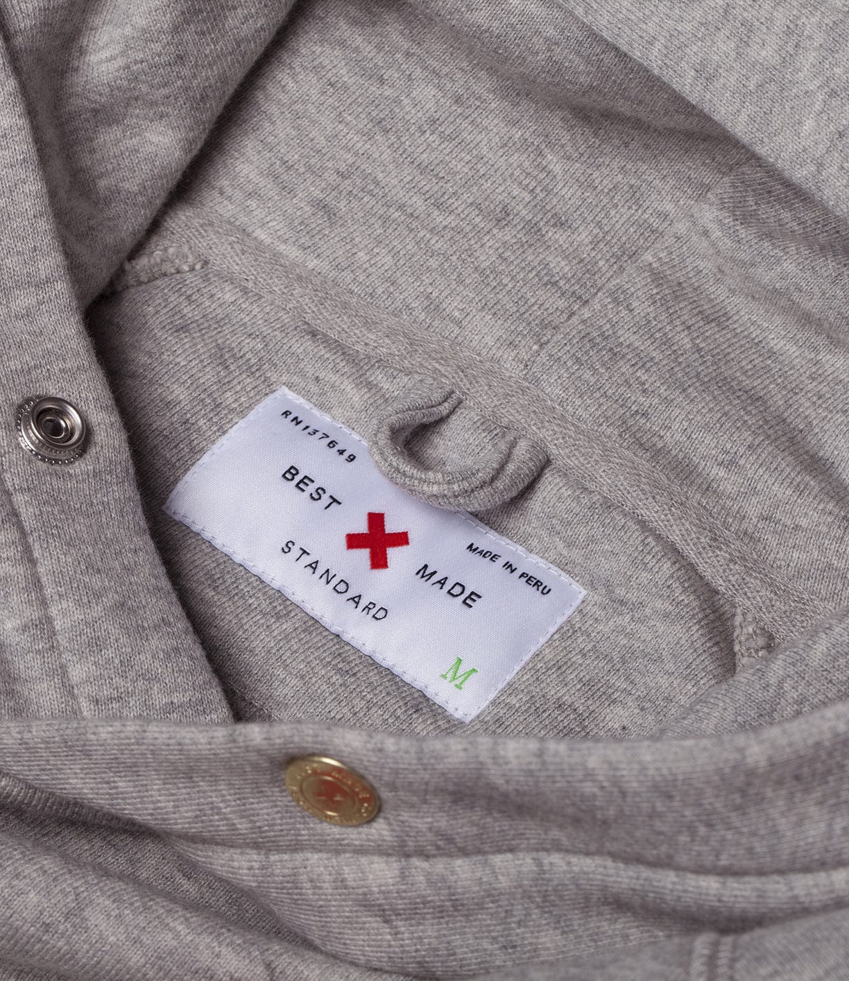 Close-up of a grey garment showing a label with Best Made and a red X logo, indicating it is made in Peru, size medium, and marked as standard.