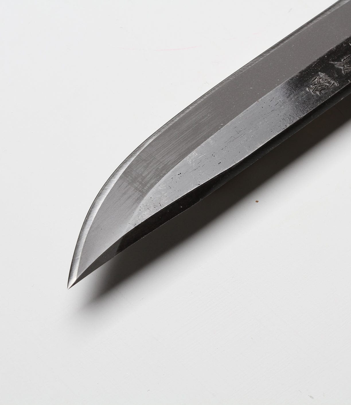 Close-up of a polished, sharp-edged knife blade, displayed on a white background. Blade has slight reflections, with a pointed tip and smooth curve.