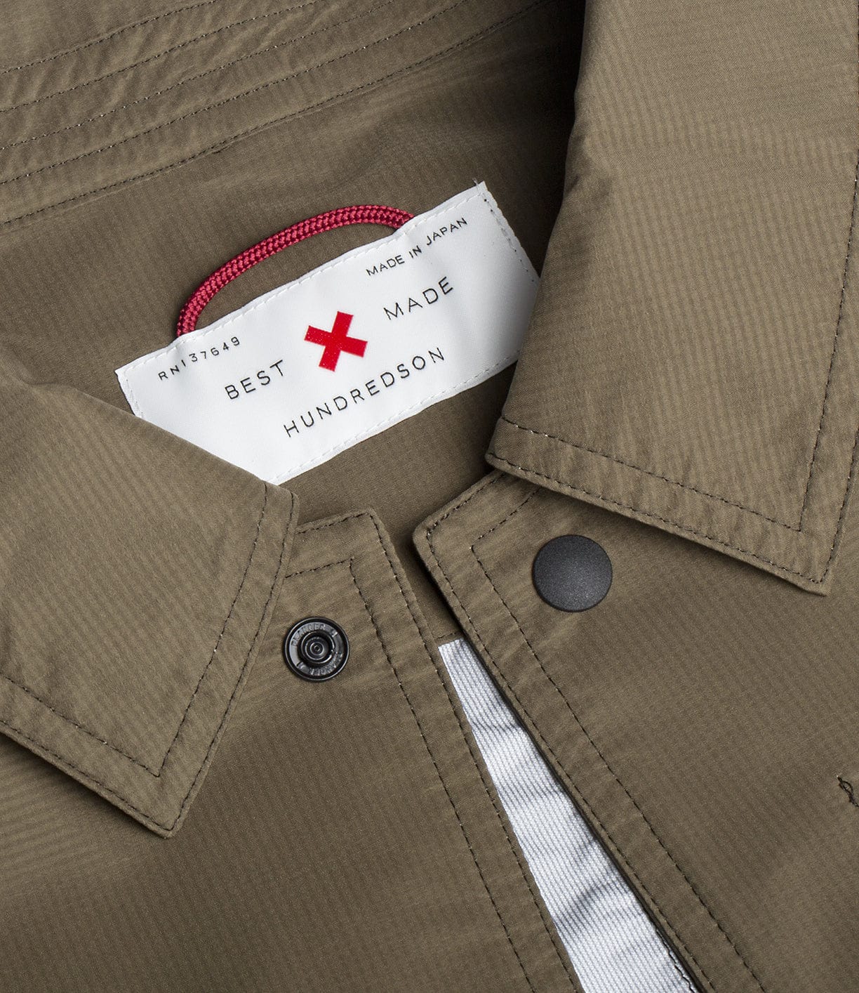 Close-up of a clothing item showing a label with the text Best Made and Hundredson. The label is affixed to a khaki fabric with visible stitching and a snap button. A red loop is also visible.