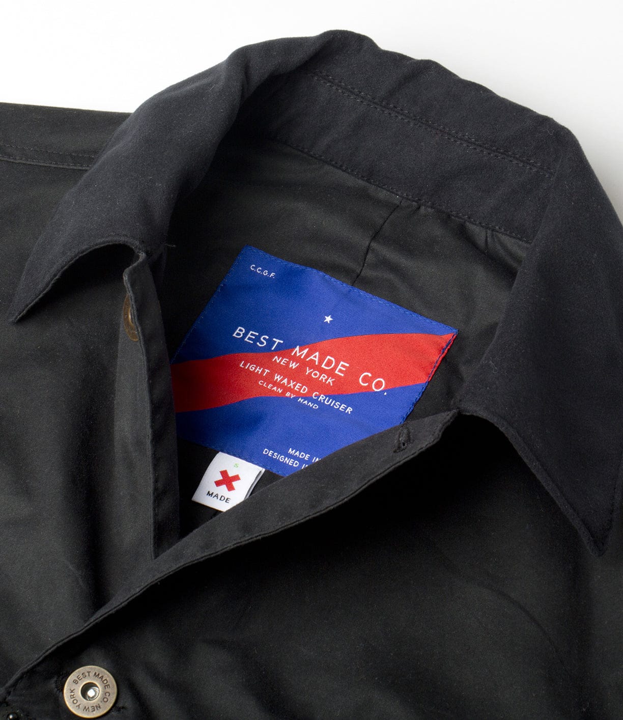 A close-up of a black jacket with a partially open collar, revealing a colorful label from Best Made Co., detailing Light Waxed Cruiser and indicating it was designed in New York and made in the USA.