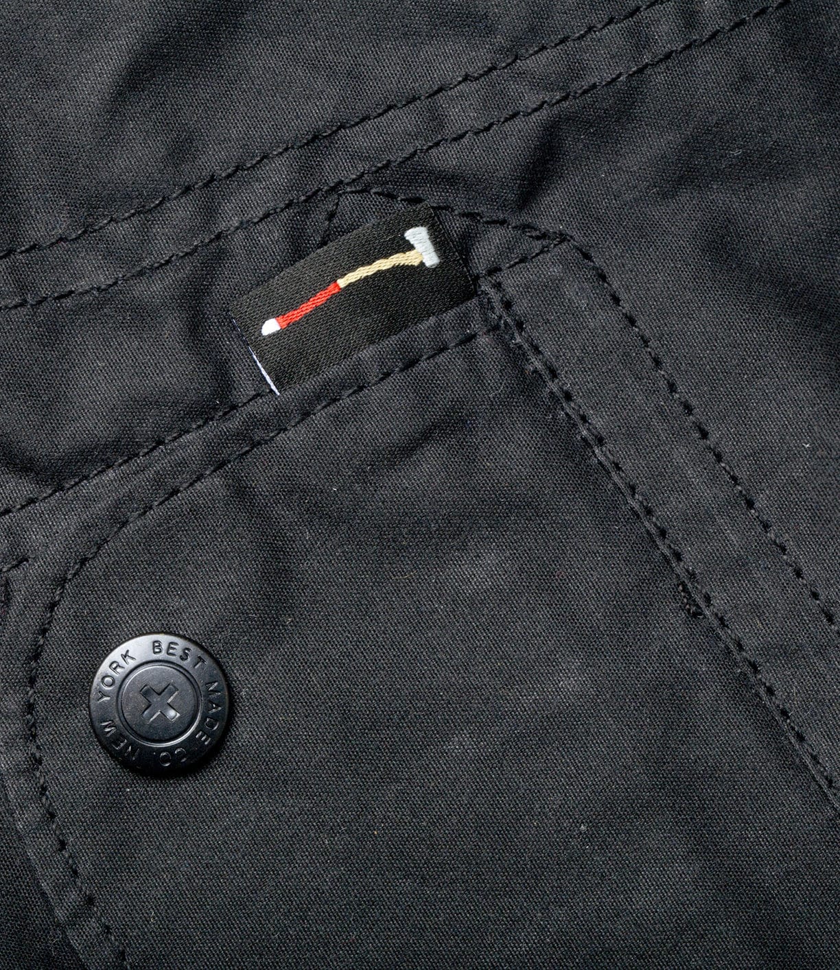 Close-up of a black fabric with a button on the left. A small black tab with red and beige stitching is partially tucked into a seam near the button.