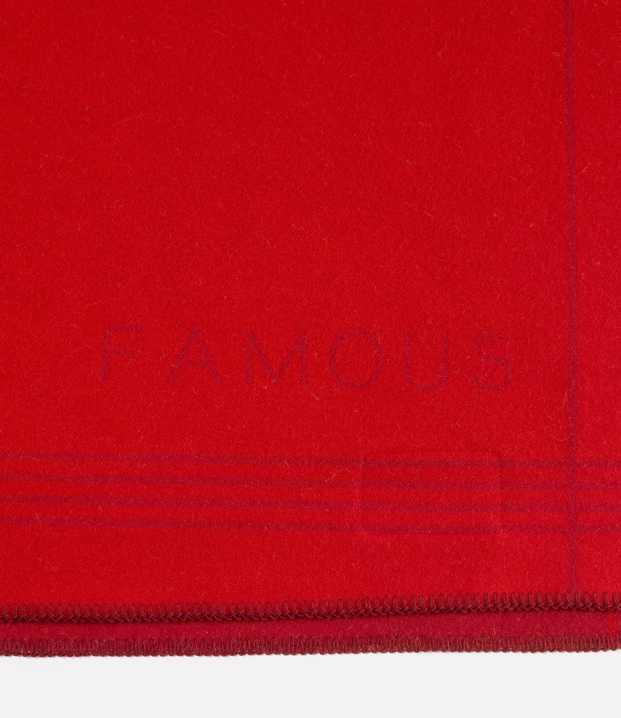 Close-up of a red fabric with the word FAMOUS subtly embossed near the bottom edge. The fabric has stitched edges and a few horizontal lines as part of the design.