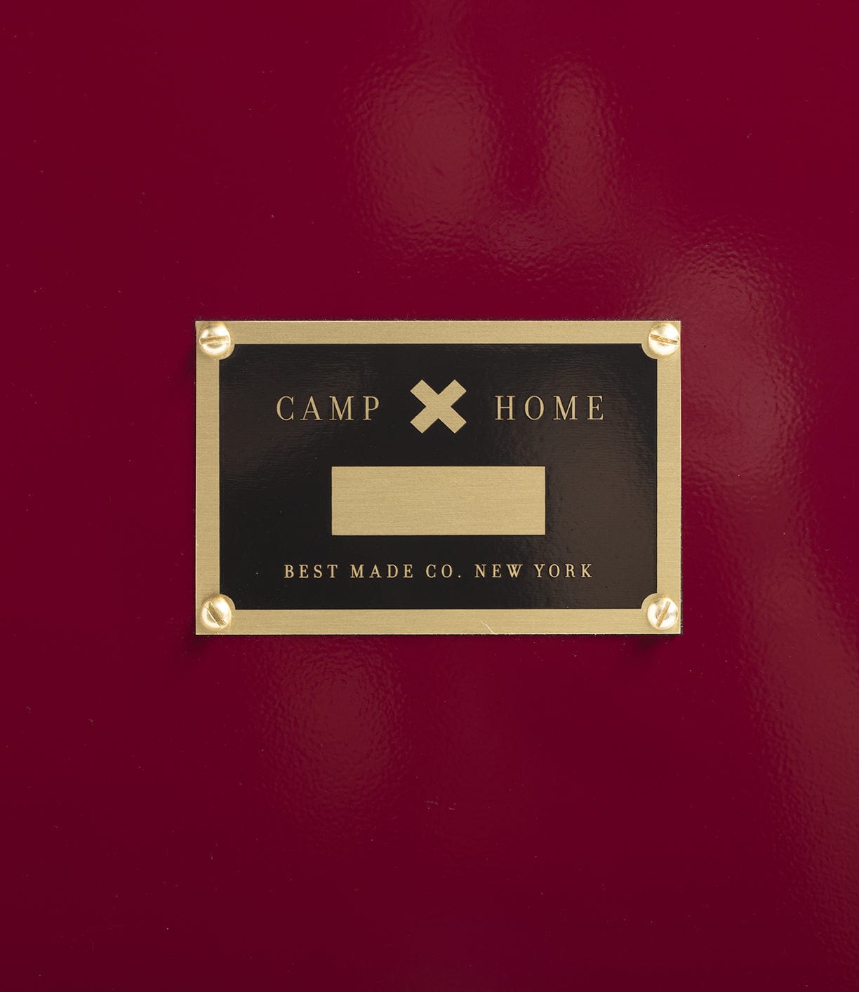 A brass nameplate with the words CAMP and HOME separated by a cross, and Best Made Co. New York beneath, is mounted on a glossy red surface.