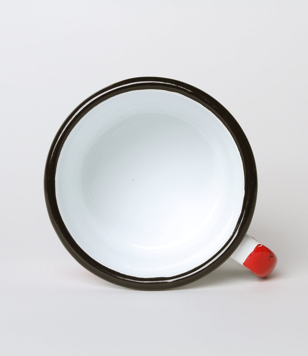 A top view of an empty, white enamel cup with a black rim and a small red and white handle placed against a plain background.