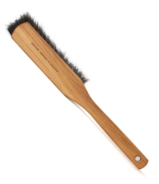 A wooden-handled cleaning brush with dark bristles. The handle has a hole at the end for hanging and is inscribed with Nature Afgor x Noquam.