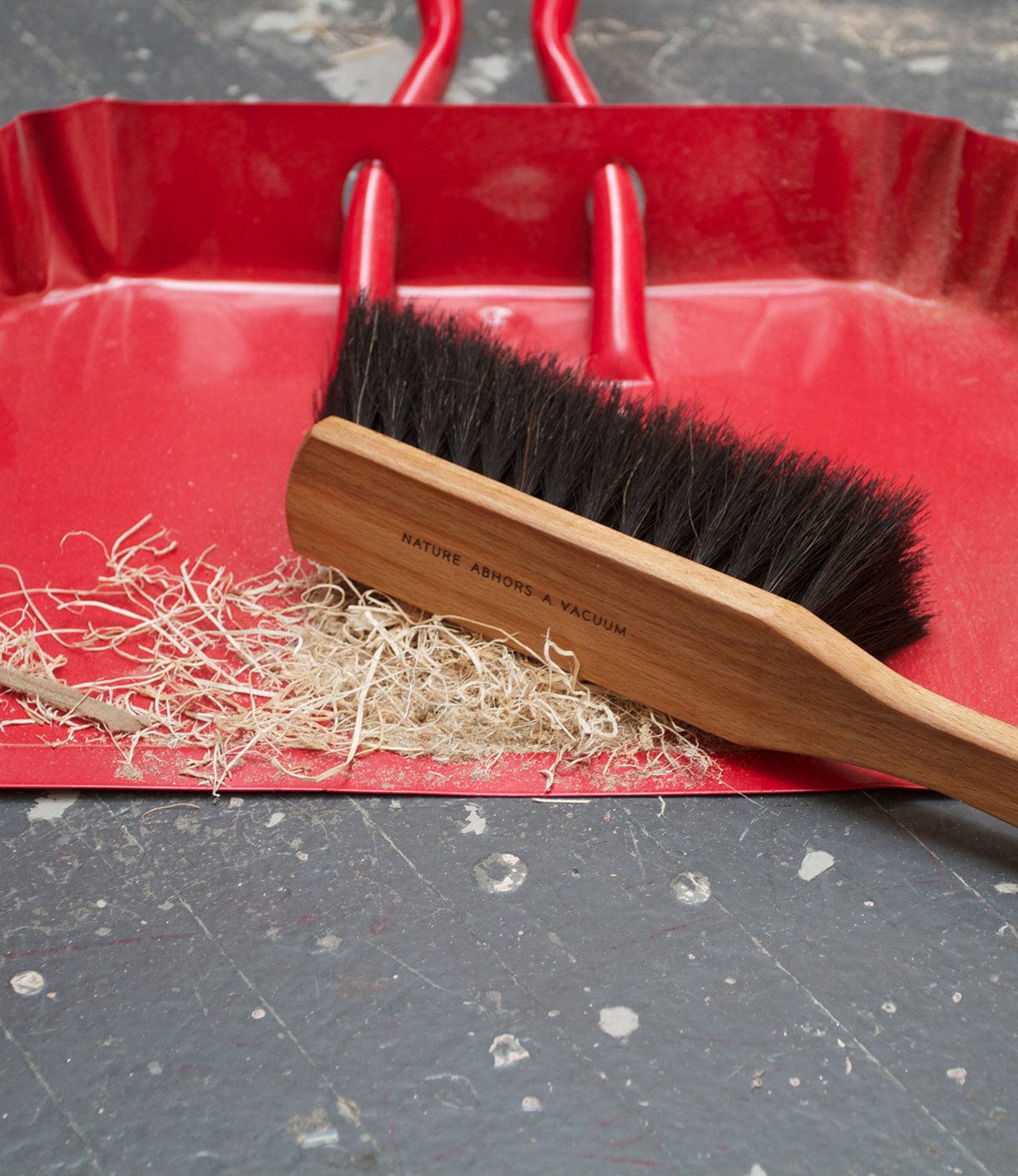 A red dustpan holding wood shavings is shown with a wooden-handled brush placed across it. The background appears to be a worn, grey surface.