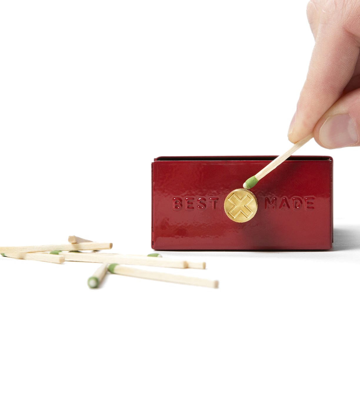 A hand striking a match against a red matchbox labeled Best Made with several unlit matches scattered nearby.