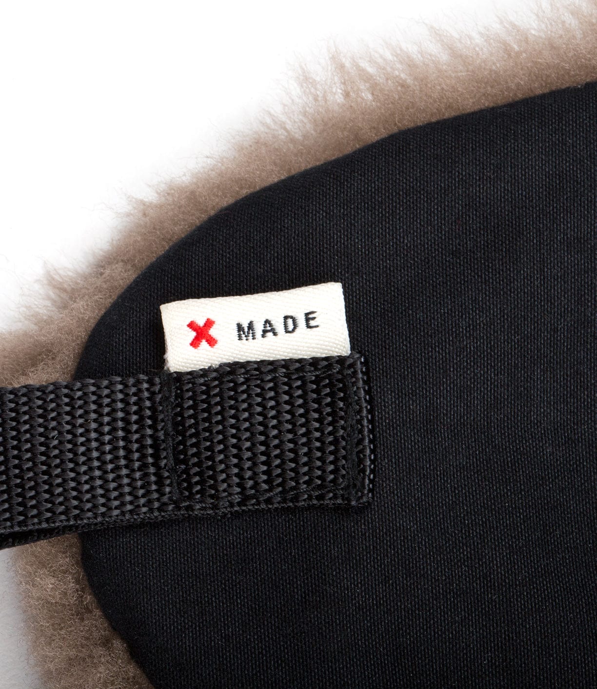 Close-up of a black fabric item with a sewn-in label reading MADE next to a red X. Black strap detail is visible on the left, with a fuzzy beige material along the edge.