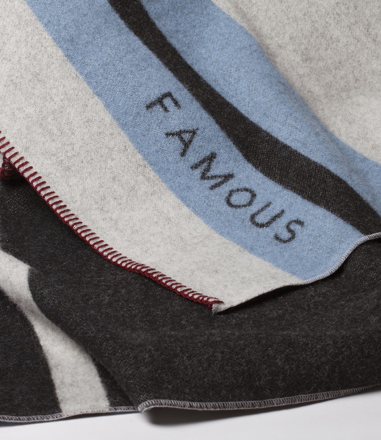 Close-up of a blanket featuring a predominantly grey and black color palette with blue stripes and the word FAMOUS written in black text. Red stitching is visible along the edge.