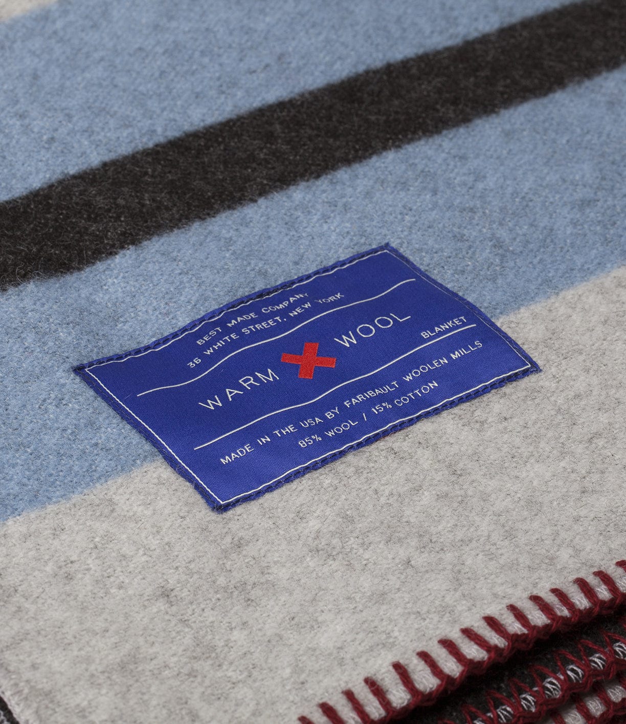 Close-up of a wool blanket with blue, gray, and black stripes. A label reads Warm Wool, listing fabric composition as 85% wool and 15% cotton, and states it's made in the USA by Farmhill Woolen Mills.