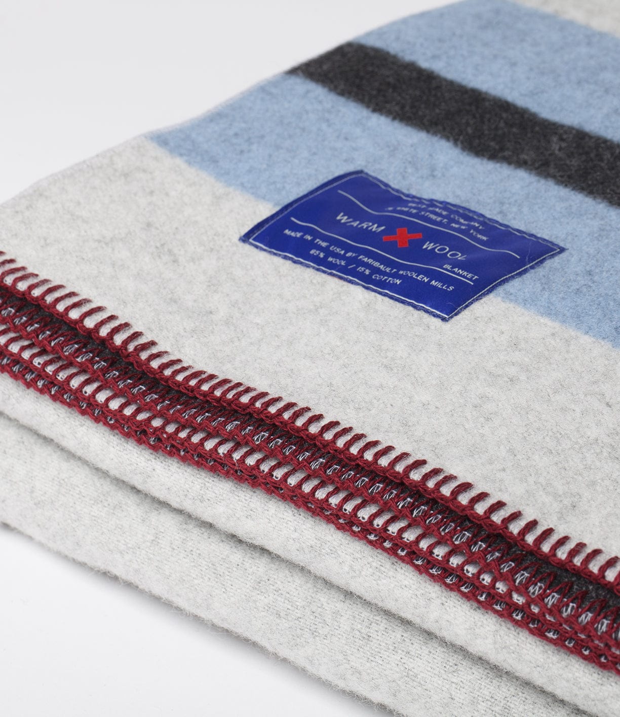 Close-up of a folded wool blanket with a blue, grey, and black stripe pattern. A blue label with red stitching reads Warm Wool and provides care instructions.