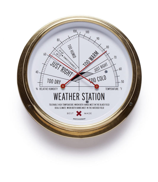 A round weather station with a brass frame, displaying humidity and temperature scales marked with Too Dry, Just Right, Too Warm, and Too Cold zones, with two red needles.