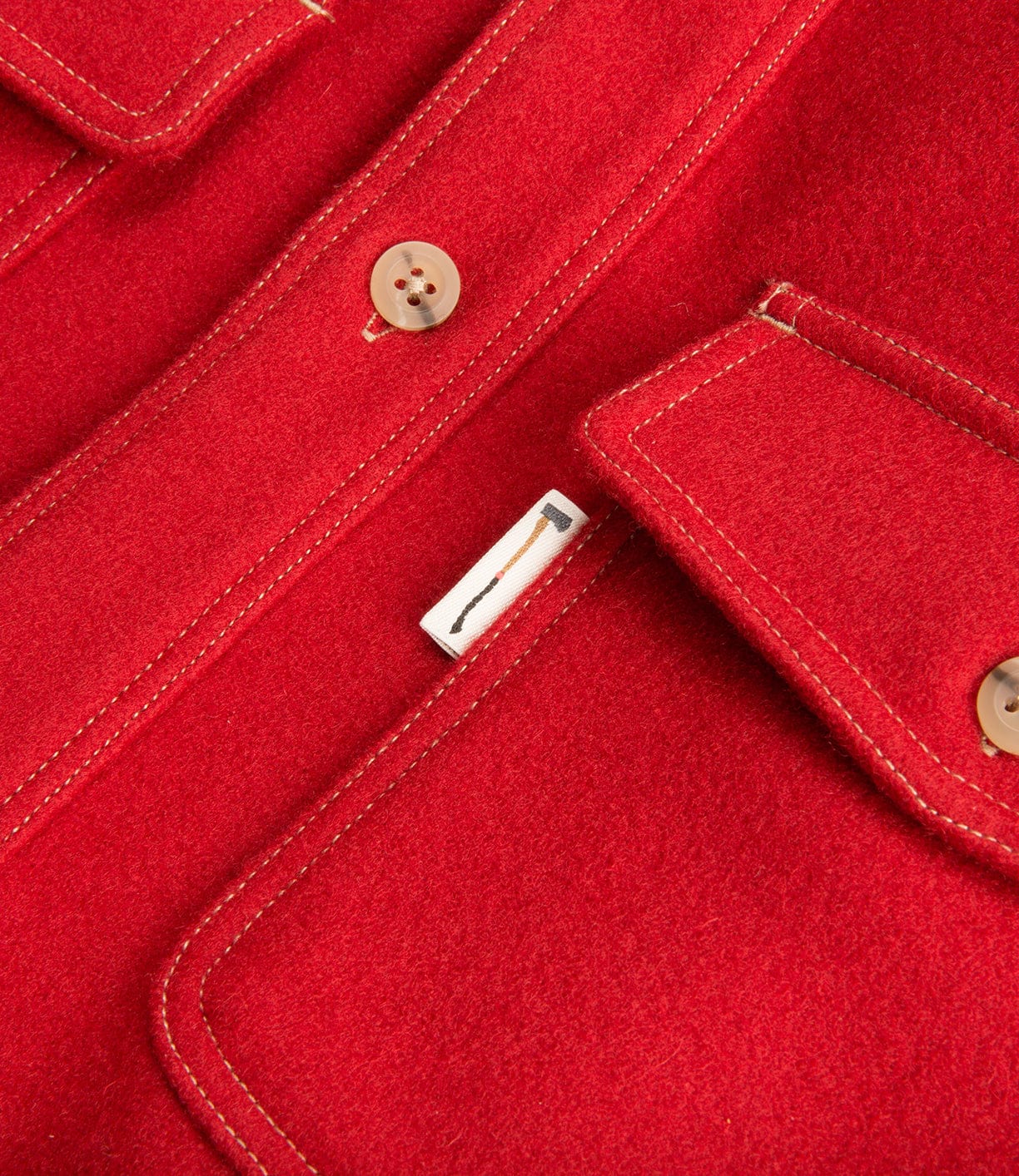 Close-up of a red wool coat featuring detailed stitching, brown buttons, and a small tag with an axe graphic on one of its pockets.