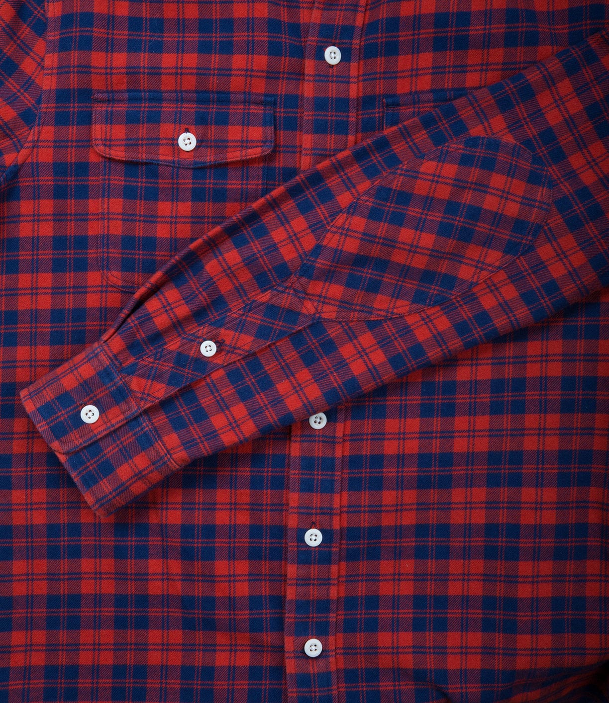 Close-up of a red and navy blue checkered flannel shirt with white buttons. The shirt's sleeve is folded across the chest.