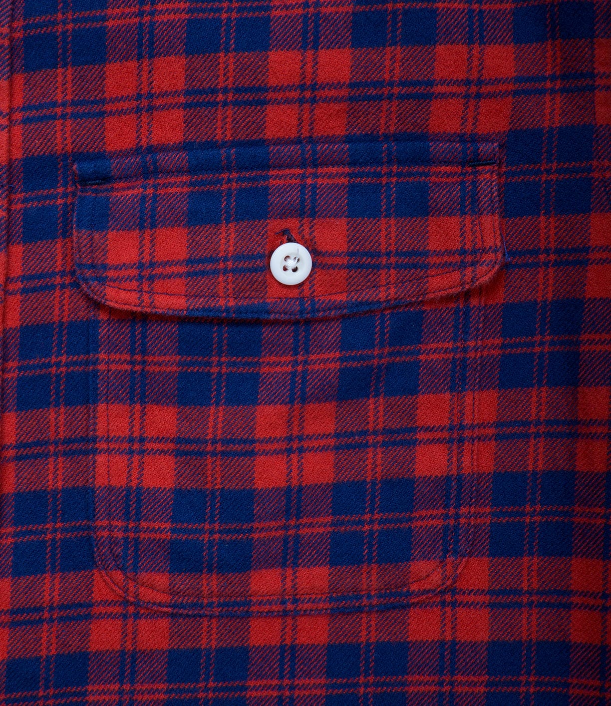 Close-up of a red and blue plaid fabric with a buttoned pocket in the center. The pocket has a white button securing the flap.